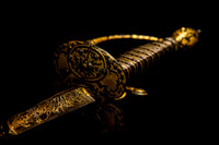 Color photograph of an ornate gilded sword with intricate gold decoration on the blade, guard and hilt against a dark background.