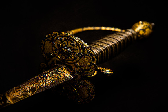 Color photograph of an ornate gilded sword with intricate gold decoration on the blade, guard and hilt against a dark background.