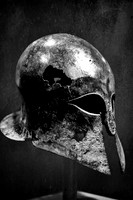 Black and white photograph of a heavily damaged ancient Greek Corinthian helmet dating from 600–550 BCE.