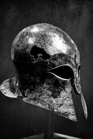 Black and white photograph of a heavily damaged ancient Greek Corinthian helmet dating from 600–550 BCE.