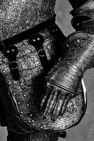 Black and white close-up photograph of ornate armor showing an articulated steel gauntlet, forearm and overlapping protection around the hip.