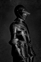 Black and white profile photograph of a historic suit of armor standing upright against a dark textured background.
