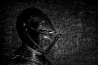 Black and white close-up profile photograph of a historic armored helmet with a narrow visor opening against a dark textured background.