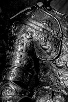 Black and white close-up photograph of highly decorated armor featuring engraved figures, faces and intricate ornamental details.
