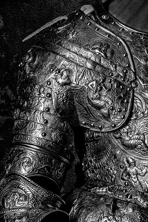Black and white close-up photograph of highly decorated armor featuring engraved figures, faces and intricate ornamental details.