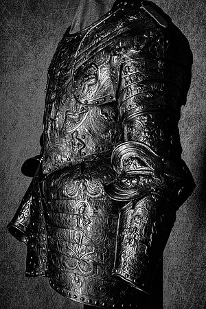 Black and white photograph of an ornate full suit of armor viewed from the side, showing its decorated breastplate, arm and overlapping leg protection.
