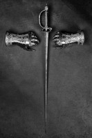 Black and white photograph of a sword positioned vertically between two armored gauntlets, arranged to suggest the shape of a cross.