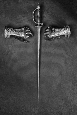 Black and white photograph of a sword positioned vertically between two armored gauntlets, arranged to suggest the shape of a cross.
