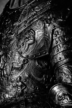 Black and white close-up photograph of highly ornate historic armor showing intricate raised decoration across the breastplate, shoulder and arm.